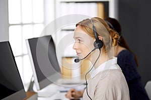 Friendly businesswoman talking by headset while sitting in office. Call center and diverse people group in business