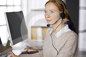 Friendly businesswoman talking by headset while sitting in office. Call center and diverse people group in business