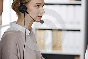 Friendly businesswoman talking by headset while sitting in office. Call center and diverse people group in business