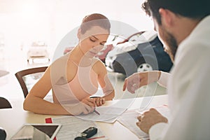 Friendly car salesman talking to a young woman and showing a new car inside showroom Signing of contract.