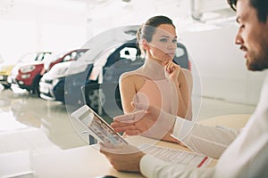 Friendly car salesman talking to a young woman and showing a new car inside showroom Signing of contract.