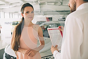 Friendly car salesman talking to a young woman and showing a new car inside showroom Signing of contract.
