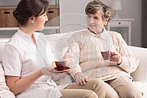 Friendly caregiver and smiling elderly woman drinking tea during meeting