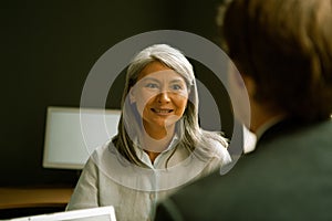 Friendly charming business woman communicates with businessman sitting with his back in foreground. Mature graying Asian