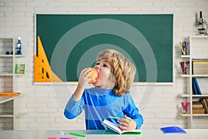 Friendly child eat apple in classroom near blackboard desk. Elementary school classroom. Kids Science education concept.