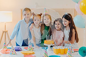 Friendly children embrace while pose near festive table, blow candles on cake, have party mood, celebrate birthday or special