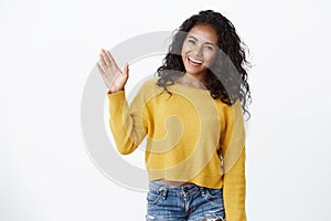 Friendly cute african-american curly-haired girl in yellow sweater raise palm and waving, saying hello, greeting guests