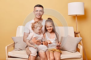 Friendly delighted happy father sitting on cozy sofa couch using smartphone with little girls daughters at home against beige wall