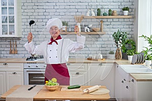 A friendly elderly chef is holding a ladle and a whisk in the kitchen.