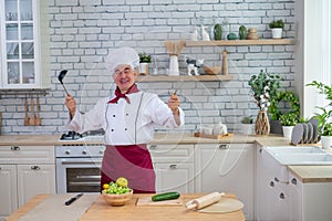 A friendly elderly chef is holding a ladle and a whisk in the kitchen.