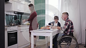 Friendly family, male father in a wheelchair in kitchen at table preparing dinner with his son and wife, talking nicely