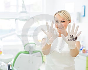 Friendly female dentist smiling and showing okay sign with fingers. Blonde doctor in gloves in the office in the medical