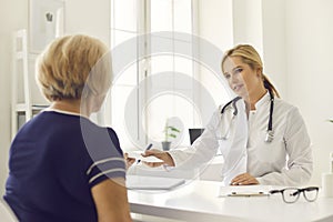 Friendly female doctor gives a patient a prescription while sitting at a table in the clinic office.