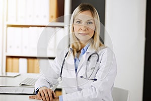 Friendly female doctor sitting in sunny clinic. Portrait of cheerful smiling physician. Medicine and healthcare concept