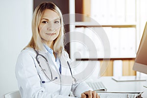 Friendly female doctor sitting in sunny clinic. Portrait of cheerful smiling physician. Medicine and healthcare concept