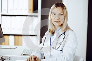 Friendly female doctor sitting in sunny clinic. Portrait of cheerful smiling physician. Medicine and healthcare concept