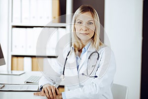 Friendly female doctor sitting in sunny clinic. Portrait of cheerful smiling physician. Medicine and healthcare concept