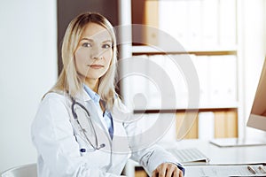 Friendly female doctor sitting in sunny clinic. Portrait of cheerful smiling physician. Medicine and healthcare concept