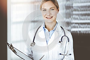 Friendly female doctor standing and holding clipboard in clinic . Portrait of cheerful smiling physician. Perfect