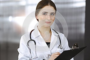 Friendly female doctor standing and holding clipboard in modern clinic. Portrait of cheerful smiling physician