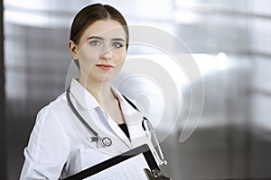 Friendly female doctor standing and holding clipboard in modern clinic. Portrait of cheerful smiling physician