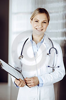 Friendly female doctor standing in sunny clinic. Portrait of cheerful smiling physician. Perfect medical service in