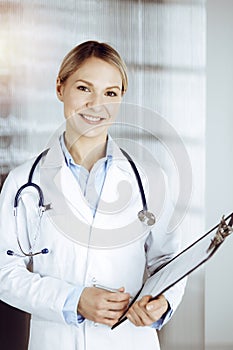 Friendly female doctor standing in sunny clinic. Portrait of cheerful smiling physician. Perfect medical service in