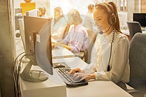 Friendly female helpline operator with headphones working on computer in call center