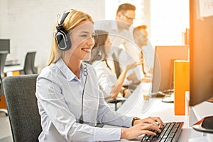 Friendly female helpline operator with headphones working on computer in office.