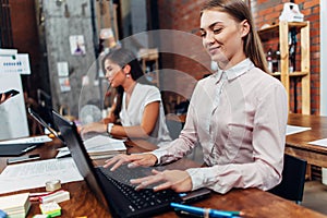 Friendly female office workers wearing formal workwear typing on laptop keyboard working in creative agency