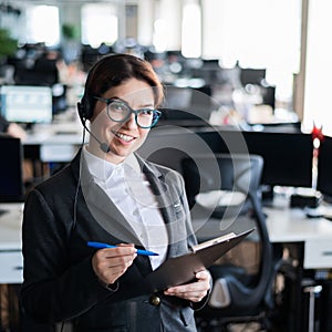 A friendly female support employee with glasses and a business suit is talking to a client on the headset. A woman with