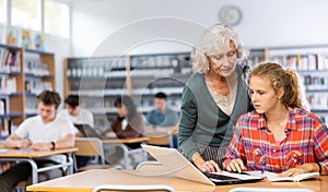 Friendly female teacher helps schoolgirl find information in laptop in school library