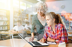 Friendly female teacher helps schoolgirl find information in laptop in school library