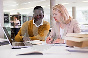 Friendly female tutor helping to diligent african-american man preparing for exam in library