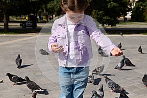 Friendly joyful little girl feeds pigeons in summer park. The concept of kindness and care for animals. Happy childhood