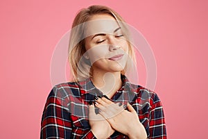 Friendly kind hearted female keeps eyes shut, hands on chest, expresses her gratitude to someone, poses against pink background. A