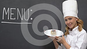 Friendly lady cook holding cake on plate, menu written in blackboard, horeca