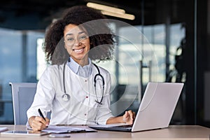 Friendly latino paediatrician in medical coat sitting by desktop with laptop and making notes by hand indoors. Happy