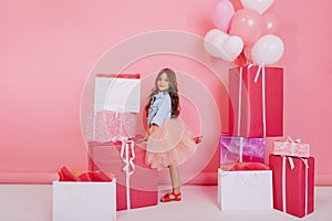 Friendly little girl with long curly hair in pink tulle skirt smiling to camera suround colorful big giftboxes, balloons