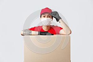 Friendly-looking asian delivery guy in red cap and t-shirt, courier service lean on cardboard box or client package