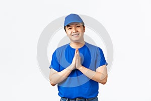 Friendly-looking handsome asian delivery man in blue uniform, holding hands in plead, palms together to say thank you