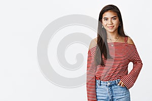 Friendly-looking stylish young indian teenage girl in striped blouse and jeans standing against gray background smiling