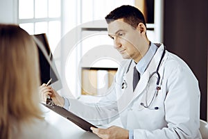 Friendly male doctor and patient woman discussing current health examination while sitting in sunny clinic. Perfect