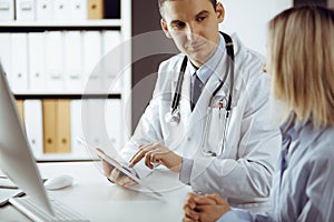 Friendly male doctor and patient woman discussing current health examination while sitting in sunny clinic. Perfect