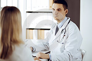 Friendly male doctor and patient woman discussing current health examination while sitting in sunny clinic. Perfect