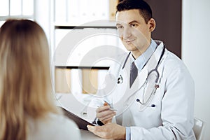 Friendly male doctor and patient woman discussing current health examination while sitting in sunny clinic. Perfect