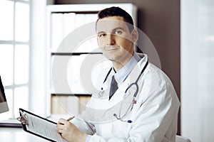 Friendly male doctor sitting and working with clipboard of medication history records in clinic at his working place