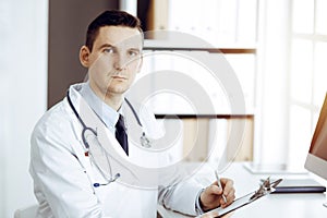 Friendly male doctor sitting and working with clipboard of medication history records in sunny clinic. Medicine and