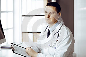 Friendly male doctor sitting and working with clipboard of medication history records in sunny clinic. Medicine and