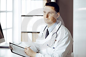 Friendly male doctor sitting and working with clipboard of medication history records in sunny clinic. Medicine and